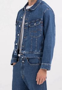Blå denimjacka med klassisk snitt, med knappframsida, bröstfickor och en blekt yta. Matchas med matchande denimbyxor.