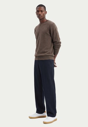 Homme portant un pull marron, un pantalon à jambes larges bleu marine et des baskets blanches, debout les mains derrière le dos sur un fond uni.