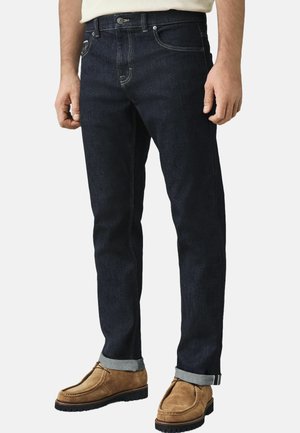 Dark blue denim jeans with a straight cut, featuring light stitching, five pockets, and a cuffed hem, paired with tan suede shoes.
