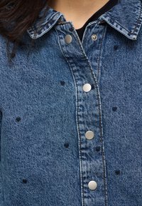 Denimjakk hõbedaste tõmblukknappide ja väikeste mustade südamekujudega, seljas musta topi kohal, näidates krae ja ülemist rinnaosa.