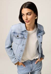 Veste en denim bleu clair avec des accents argentés éparpillés, des fermetures à boutons et un col classique, assortie à un t-shirt blanc.