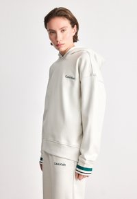 White hooded sweatshirt with green embroidered "Calvin Klein" logo. Ribbed cuffs with teal and dark blue stripes. Smooth texture, relaxed fit.