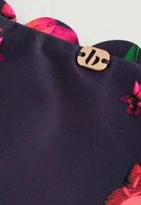 Primer plano de tela oscura con borde festoneado, estampado floral y una pequeña etiqueta dorada con una "b" minúscula, sujeta con hilo negro.