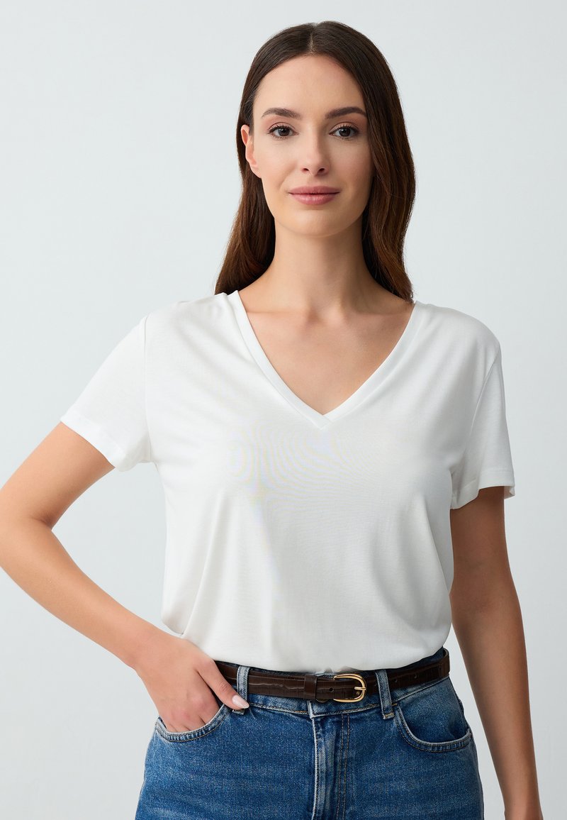 T-shirt bianca a maniche corte con scollo a V, tessuto morbido, vestibilità relaxed, abbinata a jeans di denim blu e una cintura marrone con una chiusura in oro.