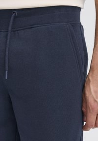 Tumesinised dressipüksid soonikvöökohaga, reguleeritava nööriga ja küljetaskutega pehmest, tekstuurse kangaga.