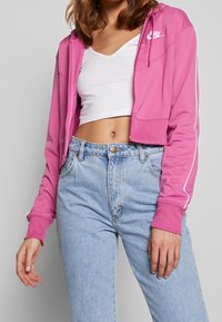 Beskarvad rosa zip-up hoodie med vita detaljer och elastiska ärmslut, bärs över en vit V-ringad topp och i kombination med högmidjade ljusblå jeans.
