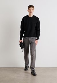 Crna dukserica s okruglim izrezom, sivi traperice i crne tenisice. Model drži crnu torbu i stoji pred jednostavnom pozadinom.