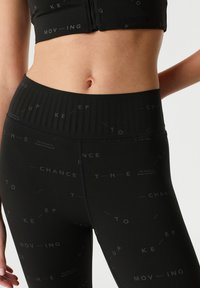 Černý sportovní crop top a legíny s vysokým pasem mají hladkou texturu s rozptýlenými šedými textovými akcenty. Pas má žebrovaný design.