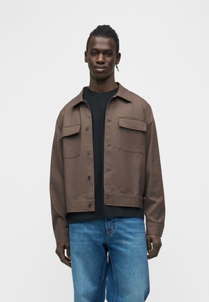 Egy férfi áll egy egyszerű háttér előtt, barna, gombos dzsekit visel fekete póló fölött és kék farmert, előre néz semleges arckifejezéssel.