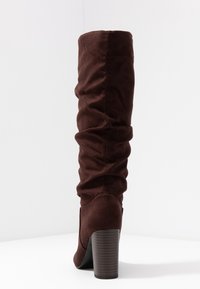 Brun mockakänga i lårhöjd med rynkad textur och klack, som visar en avsmalnande tå och synlig sömnad längs sömmen.