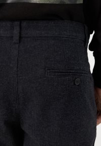 Calças texturizadas cinza-escuras com um padrão de xadrez discreto, apresentando um bolso traseiro e um detalhe em botão para fecho seguro.