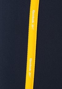 Granatowy top sportowy Reebok z wyraźnym żółtym logo oraz textured tkaniną. Akcentowany kontrastowym żółtym paskiem.