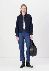 Giacca di jeans blu scuro con due tasche frontali, abbinata a jeans blu a vita alta e stivali neri alla caviglia. Il modello tiene in mano una borsa nera.
