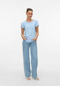 Lichtblauw, kortmouw blouse met kleine zwarte stippen, gecombineerd met wijdvallende blauwe jeans. Model staat met handen in de zakken.