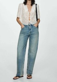 Blouse en dentelle blanche avec broderie florale et détail à nouer à l'avant, associée à un jean bleu en denim ample et des sandales noires.