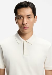 Kermansävyinen poolopaita, valmistettu teksturoidusta kankaasta. Siinä on klassinen kaulus ja kaksinappinen nappilista. Yksinkertainen ja minimalistinen muotoilu.