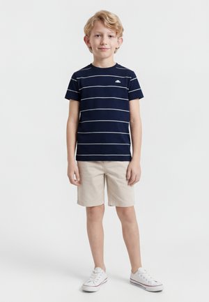 Garçon blond portant un t-shirt rayé bleu marine, un short beige et des baskets blanches, debout devant un fond blanc uni.