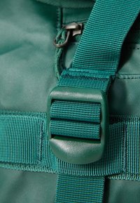 Green fabric backpack detail featuring a plastic buckle, woven strap, and a smooth zipper. Textured surface with reinforcing stitching visible.