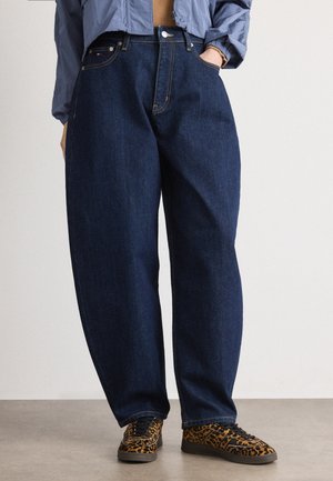 Osoba ubrana w luźne ciemnoniebieskie jeansy, szarą krótką kurtkę i trampki w panterkę stoi na szarej podłodze.