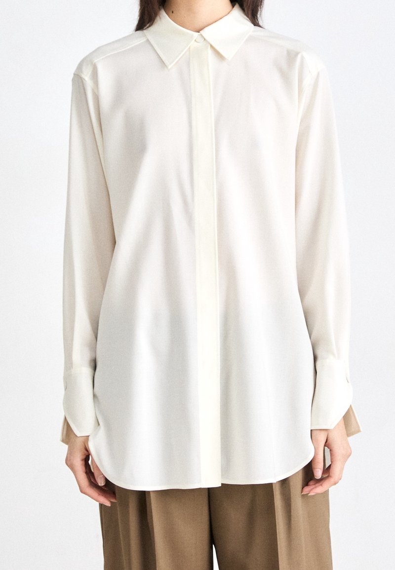 Chemise blanche à manches longues avec un col classique, devant boutonné et manches à poignets ; caractérisée par un tissu lisse, léger et une coupe décontractée.