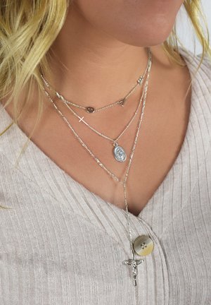 Silver layered necklaces featuring a cross, oval medal, and letter accents. Fine chains with varying lengths and textures.