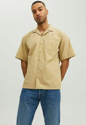 Short-sleeve, collarless shirt in light beige cotton with a front pocket and button closure, paired with blue jeans.