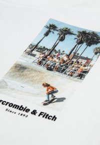 Balta kokvilnas sporta krekls ar krāsainu grafiku, kurā attēlots skeitbordists pret palmu fona, ar tekstu "Abercrombie & Fitch" apakšā.