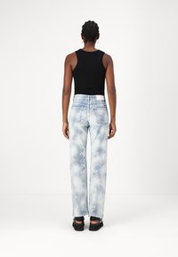 Pantalones anchos de mezclilla con patrón de serpiente en azul claro, con una cintura blanca y una camiseta sin mangas negra. Llevados con zapatos de slip-on negros, vistos desde atrás.
