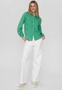 Camicetta verde a maniche lunghe con un design testurizzato, abbinata a pantaloni bianchi a gamba larga e sneakers beige, mani nelle tasche, sfondo neutro.