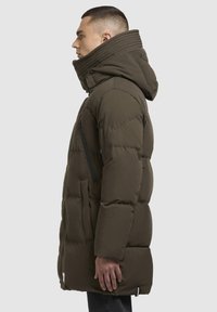 Pufjakke i mørke olivengrøn, med høj krave, sidelommer med lynlås og hætte med ribbet design. Struktureret, isoleret stof.
