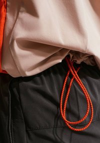 Μπεζ μπουφάν με κόκκινα και μαύρα ριγέ κορδόνια πάνω από μαύρο παντελόνι, που αναδεικνύει τις υφές του υφάσματος και τις λεπτομέρειες του ενδύματος.