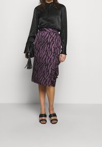 Blusa de satén negra de mangas largas, combinada con una falda morada que presenta un patrón de franjas de tigre negro. Se lleva con sandalias negras y un bolso de mano.