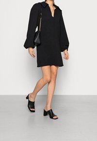 Kvinna som går i en svart långärmad minikjol med krage, svarta klackade öppen tå-mules och en svart axelremsväska med guldkedja.