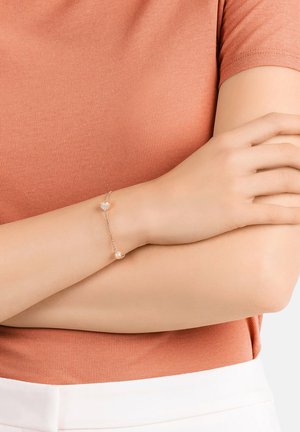 Goldenes Armband mit herzförmigen Anhängern und zwei runden Perlen, mit einem zarten Kettendesign, getragen an einem Handgelenk vor einem Hintergrund.