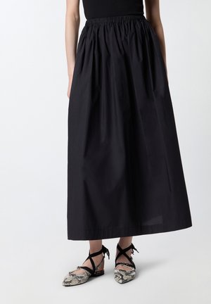 Femme portant une longue jupe noire froncée et des ballerines pointues avec des lanières croisées noires à la cheville et un motif peau de serpent.