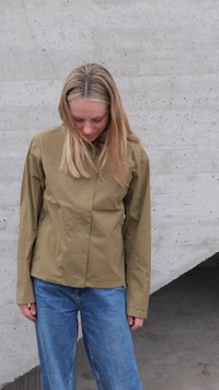 Blonde Frau in khakifarbener Jacke und blauen Jeans steht an einer Betonwand, schaut nach unten und hat die Hände neben dem Körper.