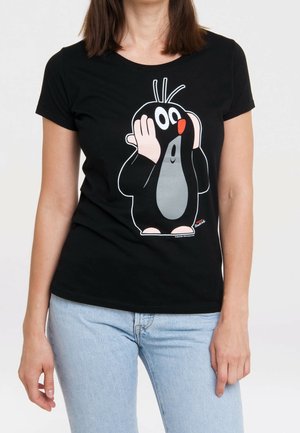 Osoba w czarnej koszulce z nadrukiem kreskówkowego kreta trzymającego twarz zaskoczeniem, ubrana w jasnoniebieskie dżinsy na jednolitym tle.