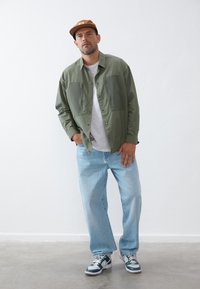 Olive green button-up shirt with two chest pockets, worn over a white t-shirt, paired with light blue loose-fit jeans and dark blue sneakers.