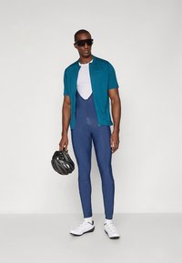 Cykelkläder inklusive en tealfärgad kortärmad tröja och marinblå långa tights, med en vit undertröja, solglasögon och en svart hjälm.