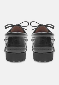 Schwarze Lederschuhe mit glänzender Oberfläche, runder Zehenform, kontrastierender strukturiertem Sohlen und Schleifenbändern, von hinten betrachtet.
