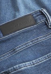 Denim tkanina v srednji modri barvi z embossed črnim usnjenim znakom "GABBA" na pasu. Ima vidno šivjenje.