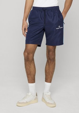 Mężczyzna ubrany w granatowe szorty Sergio Tacchini, białe skarpetki, białe trampki i białą koszulkę, stojący z rękami w kieszeniach.