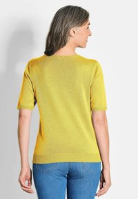 Kurzärmliger gelber Pullover mit rundem Ausschnitt und gerippten Kanten; kombiniert mit blauen Jeans. Glatte Textur, leichtes Material.