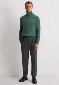 Un pull en tricot côtelé vert avec un col haut et une fermeture éclair quart, associé à un pantalon en laine gris et des chaussures à lacets noires.