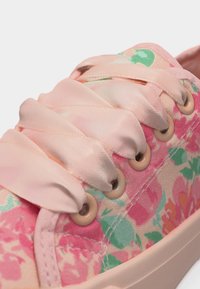 Gros plan sur une basket fleurie rose avec de larges lacets en satin passés dans des œillets beige sur une semelle blanche.