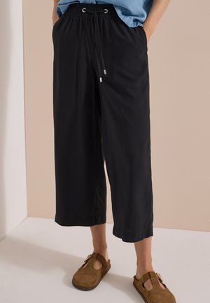 Person trägt lockere schwarze Cropped-Hosen mit Kordelzug in der Taille und braune Slip-On-Schuhe auf hellem Boden und beigem Wandhintergrund.