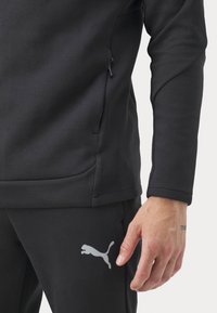 Sweat-shirt de sport noir avec manches texturées, poche zippée, et pantalon assorti arborant un logo Puma argenté réfléchissant sur la cuisse.