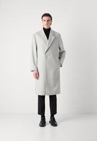 Abrigo gris claro con cuello muesca, mangas largas y bolsillos laterales. Combinado con un cuello de tortuga negro y pantalones negros.