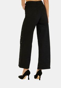 Pantaloni neri a gamba larga in tessuto strutturato, con vita alta e borchiette decorative lungo le cuciture esterne.