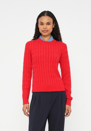 Roter gestrickter Pullover mit strukturiertem Zopfmuster. Runder Ausschnitt, gerippte Bündchen und Saum. Getragen über einem hellblauen Hemd mit Kragen.